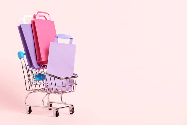 Shopping cart loaded with paper bags on a pastel pink background. Minimalist design with copy space. Concepts: market deals, seasonal sales and discounts, black friday. - Photo, image