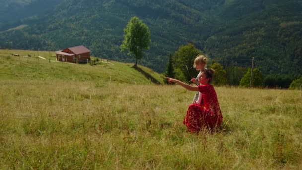 Carefree mother sitting near daughter on meadow sunny day. Happy woman relaxing with cute girl in mountains enjoying summer vacation. Young mom in red dress pointing finger at beautiful nature views. - Materiał filmowy, wideo