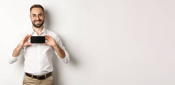 Schöner bärtiger Mann mit Brille, demonstriert Handy-Anwendung, zeigt Smartphone-Bildschirm, steht über weißem Hintergrund. - Foto, Bild