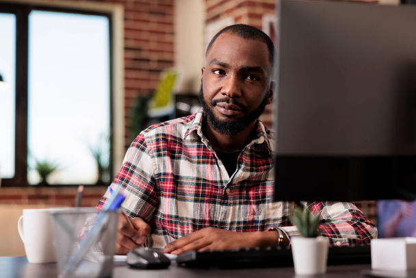 African american man taking notes on files, writing report information on notebook at office desk. Working from home on startup documents, doing paperwork and browsing internet on computer. - Foto, Bild