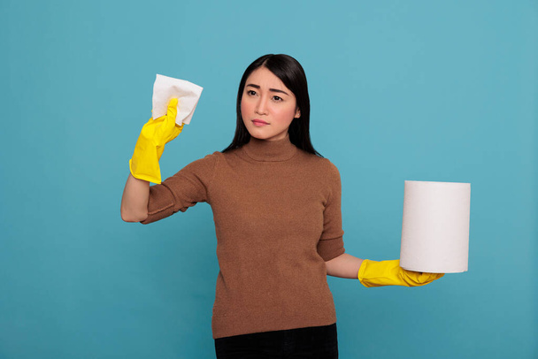 Confidence young woman with tissue paper holding in the yellow glove standing against a blue background, Cleaning home concept, Faithful satisfied female from day to day chores - 写真・画像