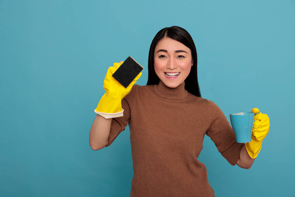 Excited smiling asian young housemaid wearing yellow gloves holding a sponge and coffee cup Cleaning home concept, Refreshment tea at break time, Woman feeling cheerful satisfied and positive - Foto, Bild