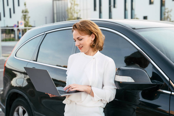 Улыбающаяся уверенная деловая женщина, использующая ноутбук, работающий онлайн, стоящий рядом с машиной. Успешный бизнес    - Фото, изображение