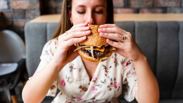 Бургери їдять дівчину. Голодна кавказька жінка їсть смачний гамбургер. Концепція нездорової їжі
 - Фото, зображення