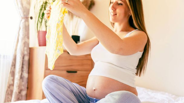 妊娠中のバッグベビー服。黄色の赤ん坊の服を保持する幸せな若い妊婦。マザーパッキングスーツケース、妊婦病院用バッグ。妊娠・産前・出産への期待 - 写真・画像
