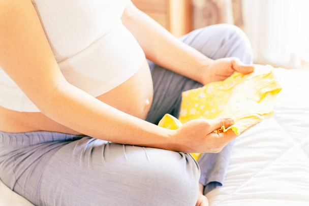 Vauvanvaatteet raskaana. Kaunis raskaus äiti keltainen vauvan vaatteita. Raskaana oleva nainen halailee vatsaa ja pakkaa äitiyssairaalan laukkua. Käsitys äitiys, raskaus, synnytys - Valokuva, kuva