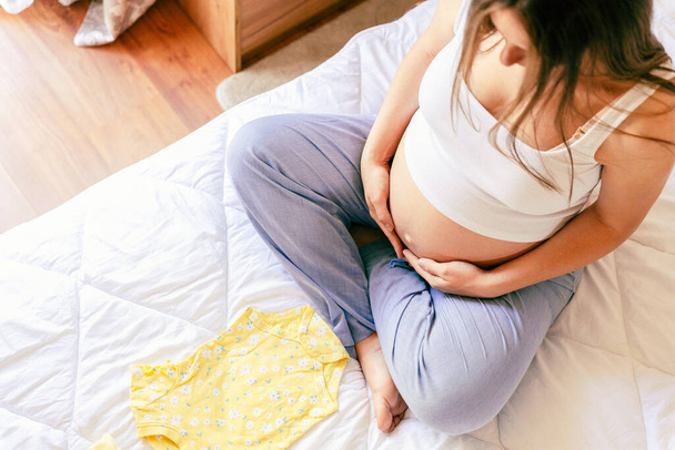 Roupas de bebê mulher grávida. Bela mãe gravidez com roupas de bebê amarelo. Mulher grávida abraçando barriga e saco de maternidade de embalagem. Conceito maternidade, gravidez, parto - Foto, Imagem