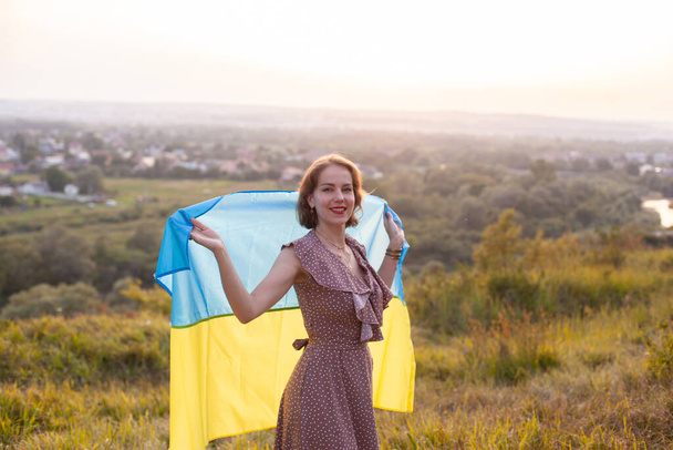 Happy Woman in long dress holding a yellow and blue flag of Ukraine fluttering in the wind on a background of sunset. Ukrainians against the war. Independence Day. Flag Day. Constitution day.  - Foto, imagen