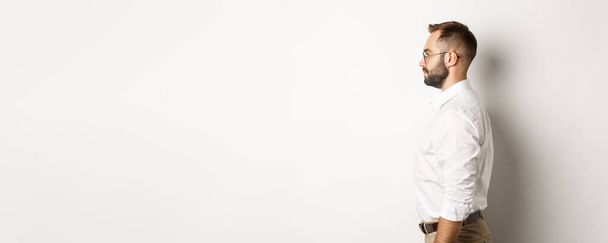 Профиль молодого бизнесмена в белой воротничковой рубашке и бежевых брюках, смотрящего влево, стоящего на фоне студии. - Фото, изображение
