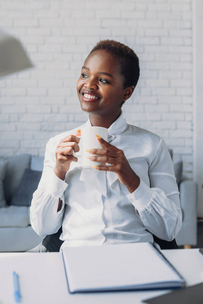 Dreamy african businesswoman enjoying coffee cup, having rest after work. Thinking over successful project, future profit, smiling, laughing, feeling joy - 写真・画像