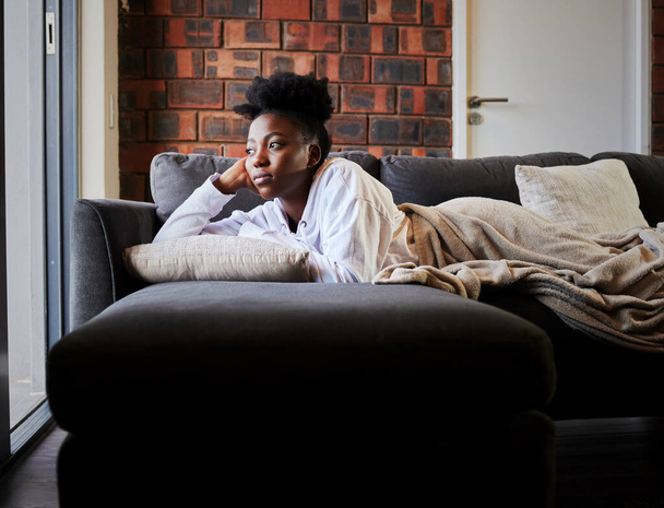Bored, depression and isolation with black woman lying on her sofa during lockdown, quarantine or pandemic. Mental health, anxiety and stress with young female thinking and looking out her window. - Fotoğraf, Görsel