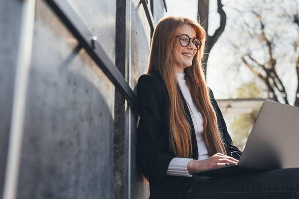 Side view of a businesswoman wearing eyeglasses working outdoors on her laptop enjoying the fresh air sitting on a bench. The concept of an open-air office - Photo, image