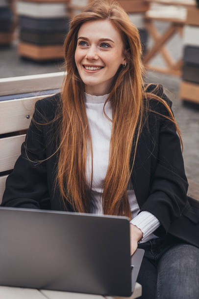 Smiling businesswoman working on pc in the fresh air sitting outside on a bench in the city park. Beautiful young girl. Modern technology. Business portrait - Foto, afbeelding