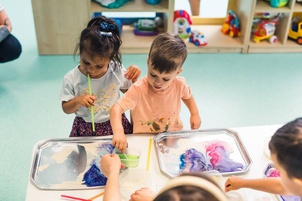 Sensory play at multi-cultural nursery school. Toddlers with their teacher playing with striped straws and milk painting, using food coloring, milk, watercolor paper, and trays. Creative kids activity - Fotoğraf, Görsel
