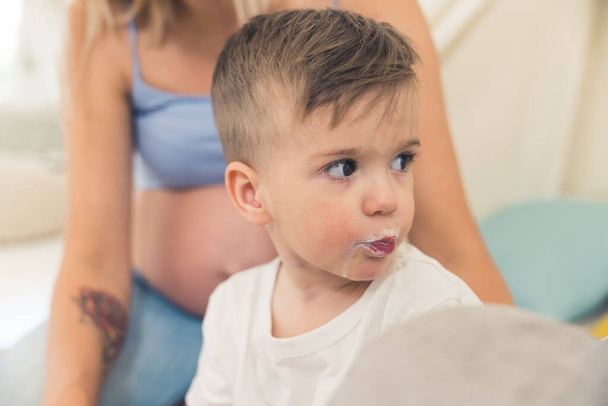 Indoor portrait closeup shot of young preschooler dark-haired boy with mouth full of milk looking back. Caucasian pregnant mother with nude big belly in the blurred background. High quality photo - Foto, imagen