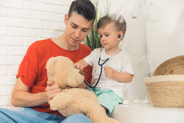 Different games with children. Medium closeup indoor shot of caucasian millennial dad with his preschooler son playing doctor with teddy bear. Stethoscope. High quality photo - Foto, afbeelding