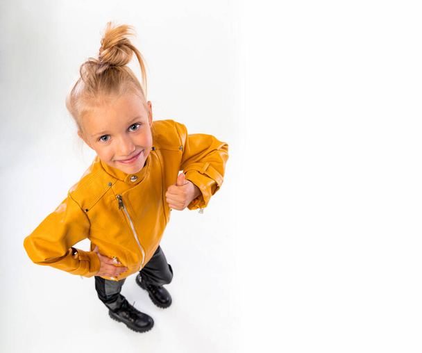 Thumbs up. Pretty blonde little girl in a leather mustard jacket, black snake texture leggings and boots. High fashion, full length standing pose, isolated against a studio background - Fotoğraf, Görsel