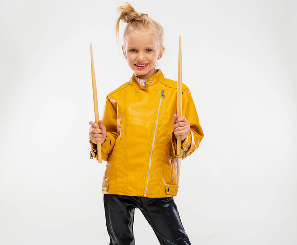Rock and roll music concept. Girl musician, holding drum sticks in hands. Dressed in a leather mustard jacket, black leggings and boots. High fashion, isolated against a studio background - Foto, imagen