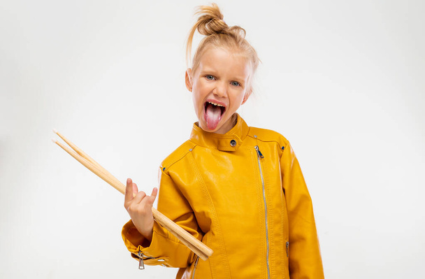 Rock and roll music concept. Girl musician, holding drum sticks in hands and shows her tongue. Dressed in a leather mustard jacket, black leggings and boots. Full length, istudio background - 写真・画像