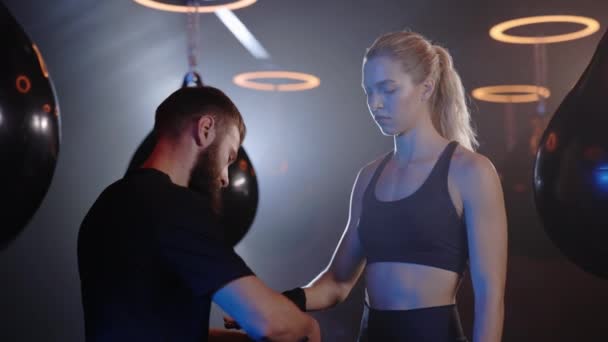 Male trainer is helping female boxer with the hand bandage on the smoky boxing gym background. Preparation for training. People and sport concept - Imágenes, Vídeo