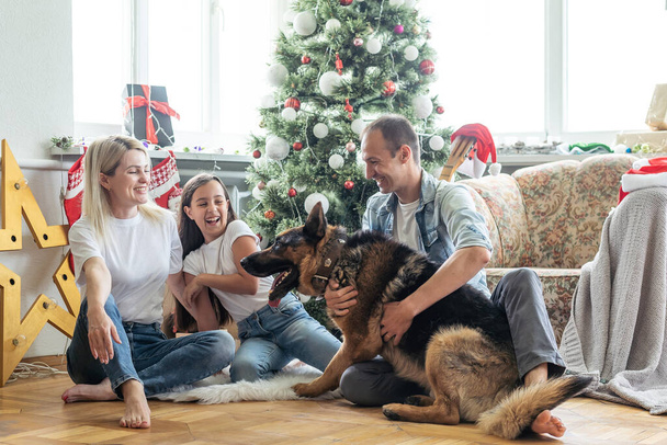 Ystävällinen perhe leikkii koiran kanssa joulukuusen lähellä. He istuvat ja nauravat.
. - Valokuva, kuva