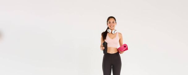 Yoga derslerine giden Asyalı güzel bir kız, kulaklıklı ve su şişeli bir bayan sporcu spor salonu eğitimi için lastik paspas taşırken gülümsüyor.. - Fotoğraf, Görsel