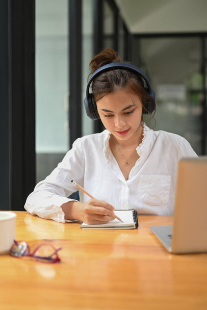 Concentrated female student wearing headphone, listening lecture during study online on laptop and making notes on notebook. - Foto, Imagem