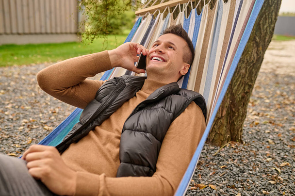 Smiling joyful attractive young Caucasian man lying in the hammock while talking on the smartphone - Фото, изображение