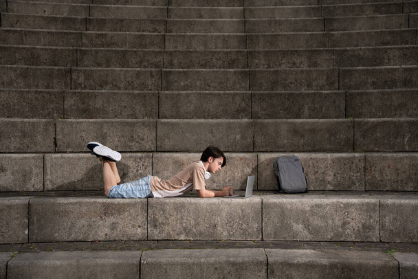 Teenager boy laying on staircase and working on laptop with dramatic lighting. - Foto, Imagen