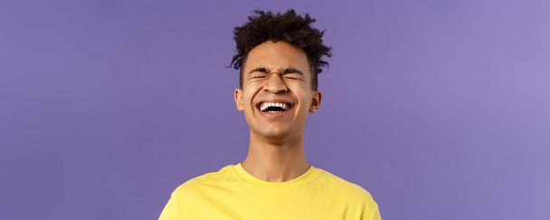 Κοντινό πλάνο πορτρέτο του χαρούμενου ανέμελου νεαρού που γελάει δυνατά, γελώντας πάνω από ένα αστείο, σκύβοντας προς τα πίσω και κλείνοντας τα μάτια ενώ χαζογελάει πάνω από μια αστεία ταινία, μωβ φόντο. - Φωτογραφία, εικόνα