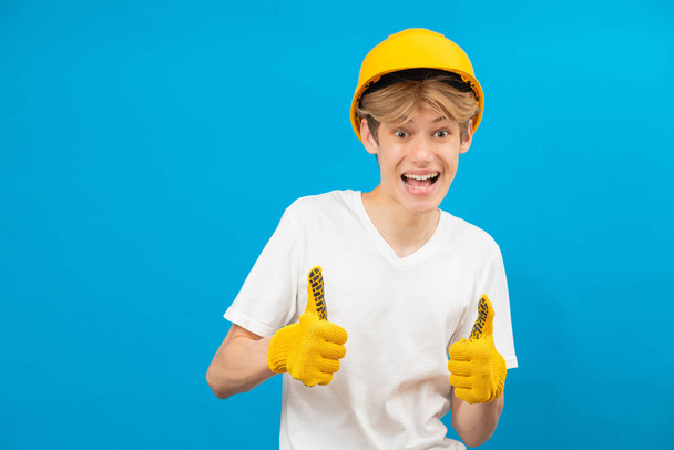 Happy handsome teen builder in white t-shirt and gloves in his hand with yellow helmet standing in studio on blue background. Caucasian craftsman isolated raising both thumbs up, smiling and confident. - Foto, imagen