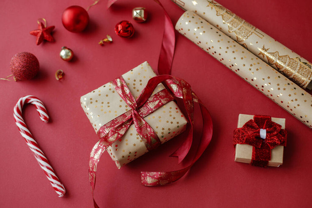 Christmas composition. Stylish christmas gift, golden wrapping paper, ribbon and festive decorations on red background. Wrapping christmas gifts concept. Happy Holidays ! - Photo, image