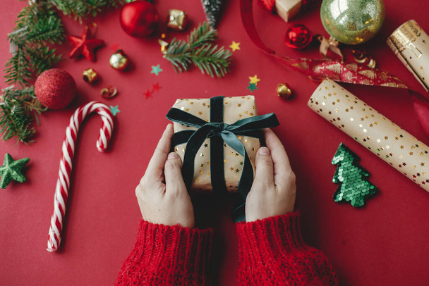 Joyeux Noël ! Mains tenant cadeau de Noël élégant, papier d'emballage doré, ruban et décorations festives sur fond rouge. Salutations de saison. Joyeuses fêtes - Photo, image