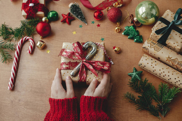 Kezek díszítő stílusos karácsonyi ajándék és arany papír, piros szalag és ünnepi dekorációk rusztikus fa asztal. Boldog karácsonyt és boldog ünnepeket! Üdvözlet az évszakoknak - Fotó, kép