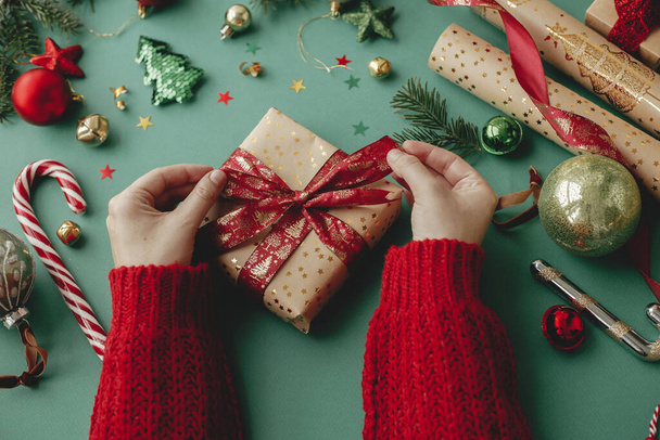 Wesołych Świąt! Ręce zawijania stylowy prezent na Boże Narodzenie, złoty papier pakowy, czerwona wstążka i świąteczne dekoracje na zielonym tle. Pakowanie prezentów świątecznych. Wesołych Świąt - Zdjęcie, obraz