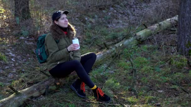Beautiful girl with a backpack drinks tea from a thermos in the forest sitting on a log. Woman warming up by drinking hot drink. Female traveler sitting and relaxing in nature in spring among trees - Video, Çekim