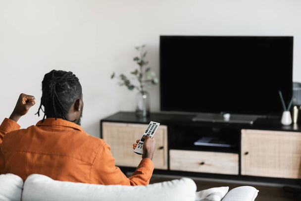 African american man cheerfing for favourite team, watching sports game on TV, celebrating victory, sitting on sofa and looking at television set with empty screen - Foto, Bild