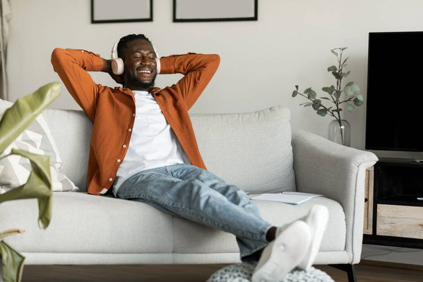 Calm african american man resting on couch, listening to music, audio book, podcast, enjoying meditation for peaceful mind in wireless headphones, leaning back, copy space - Photo, image