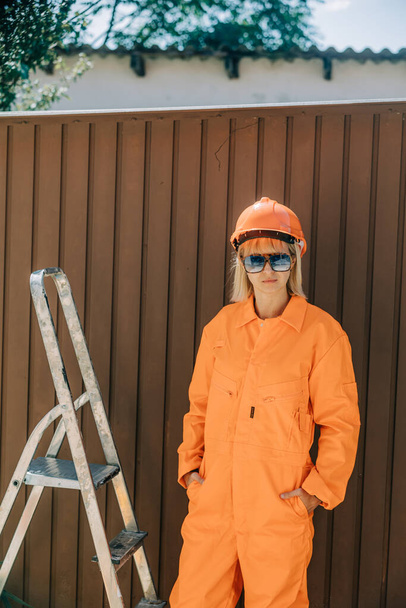 Stylish woman in orange suit, hardhat and sunglasses next to stairs on brown background - Photo, Image