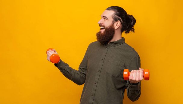 Strong cheerful bearded man lifts up easy dumbbells in a yellow studio while looking aside. - Foto, imagen