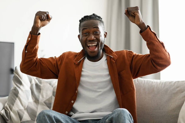 Overjoyed african american man screaming in excitement and gesturing YES, sitting on sofa in living room at home. Triumphant black guy shouting WOW, cheering for sports team on TV - Zdjęcie, obraz