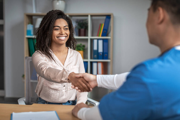 Smiling young african american woman patient shaking hands with mature asian male doctor in clinic office interior. Excellent medical service, examination with therapist and meeting for health care - Foto, immagini