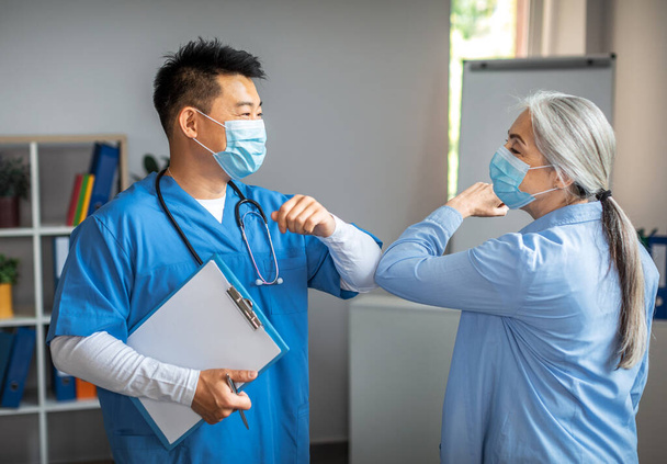 Smiling adult chinese man doctor in protective mask greets with elbows to old woman patient in clinic office interior. Meeting during covid-19 pandemic, treatment with therapist, medical health care - 写真・画像