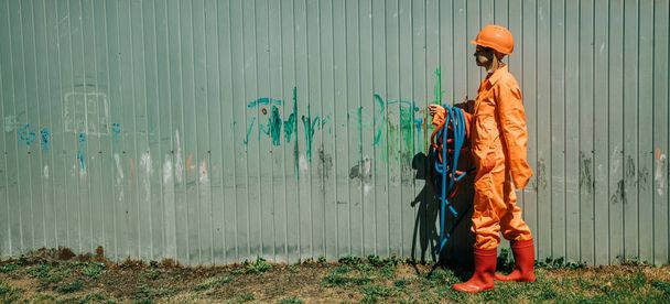 Stylish woman in orange suit, hardhat, pipes and sunglasses on gray background - 写真・画像