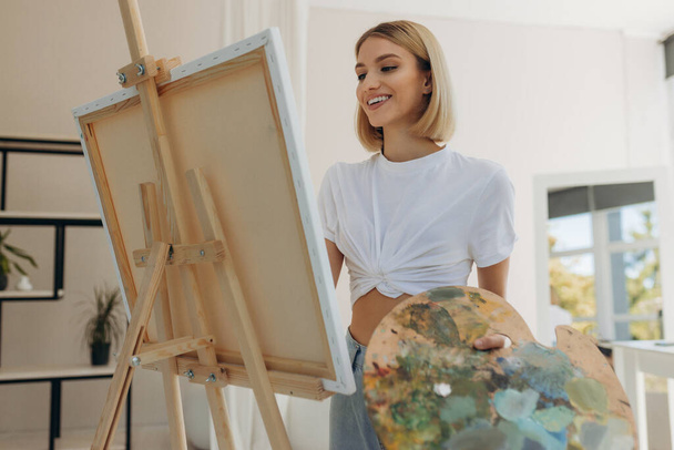 The artist paints in the studio. Attractive girl wearing a white T-shirt - Φωτογραφία, εικόνα