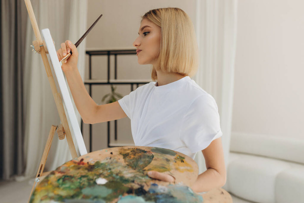 The artist paints in the studio. Attractive girl wearing a white T-shirt - Φωτογραφία, εικόνα