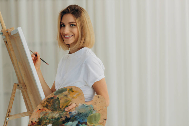 Positive young woman holding paintbrush and palette with colorful paints near easel. Artist - 写真・画像