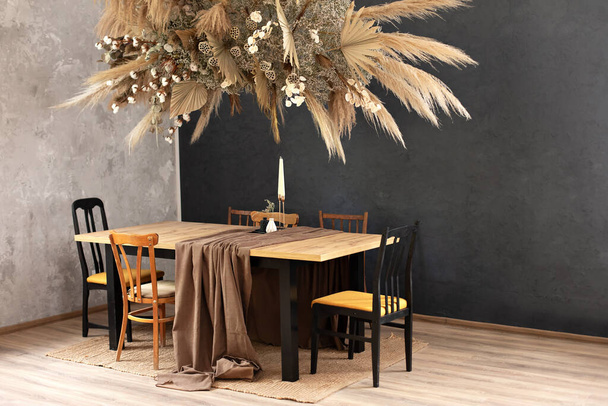 木製のテーブルと椅子付きのスタイリッシュなダイニングルーム。休日の家族の夕食。家具付きのキッチンインテリア。リビングルームの壁に組成乾燥した装飾植物。秋の装飾が施された居心地の良い部屋 - 写真・画像