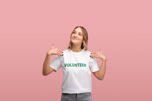 Fröhliche junge Frau schaut lächelnd weg und demonstriert Freiwilligenaufschrift auf weißem T-Shirt vor rosa Hintergrund - Foto, Bild