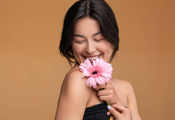 Merry young Asian female virgin smiling with closed eyes and touching shoulder with fresh gerbera flower against brown background - Photo, Image